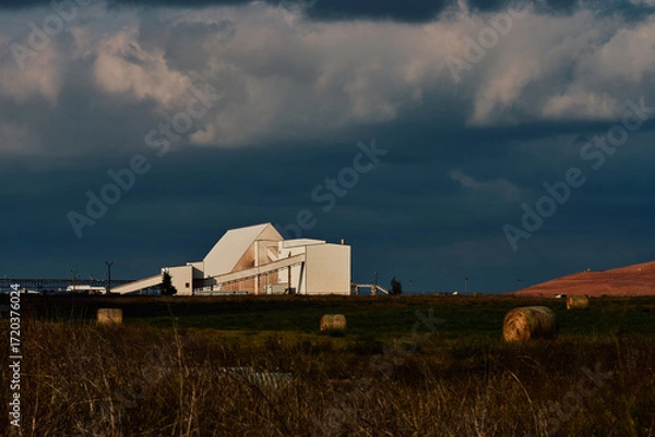 Fototapeta Industrial potash facility in Saskatchewan, Canada, with white processing buildings, conveyor systems, hay bales in the foreground, and tailings pile under dramatic cloudy sky.