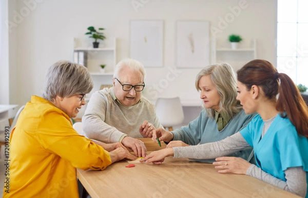Fototapeta Happy smiling senior elderly people group sitting at table in retirement home collecting geometric figures with young friendly nurse. Dementia prevention and leisure time in nursing home concept.