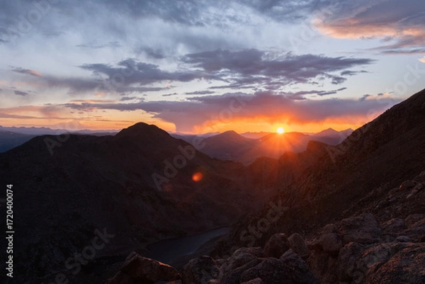 Fototapeta Glowing evening sun setting behind rugged mountain range with colorful clouds, tranquil natural scene in golden light