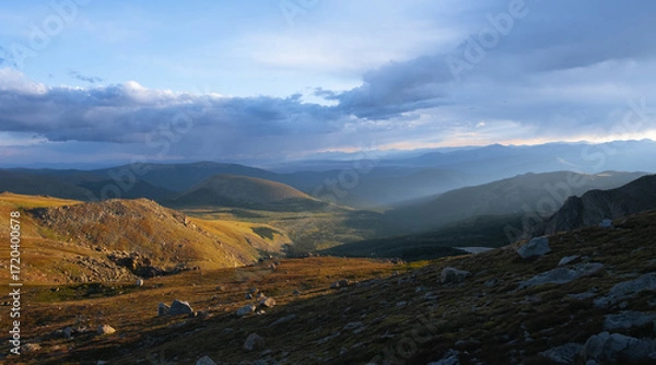 Fototapeta Sun rays breaking through clouds over mountain valley, scenic wilderness landscape with rolling hills and dramatic light