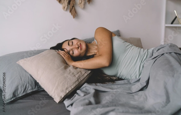 Fototapeta Brunette woman enjoying restful sleep at home, arms folded under pillow, gentle smile on face, representing emotional security, slow living and balance in everyday routine.