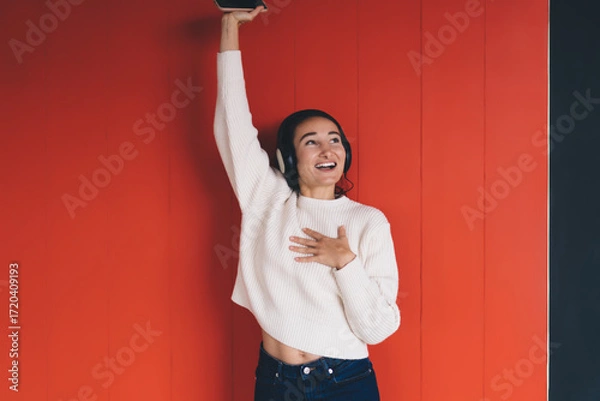 Fototapeta Young happy woman enjoying music in headphones, raising phone, smiling with joy against red wall, symbolizing connection, entertainment, and emotional expression.