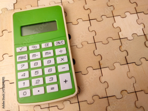 Fototapeta Closeup a green calculator resting on a surface covered with wooden jigsaw puzzle pieces