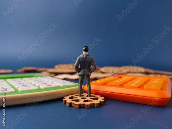 Fototapeta The image depicts a miniature figure of a businessman standing on a wooden gear, with calculators in the foreground
