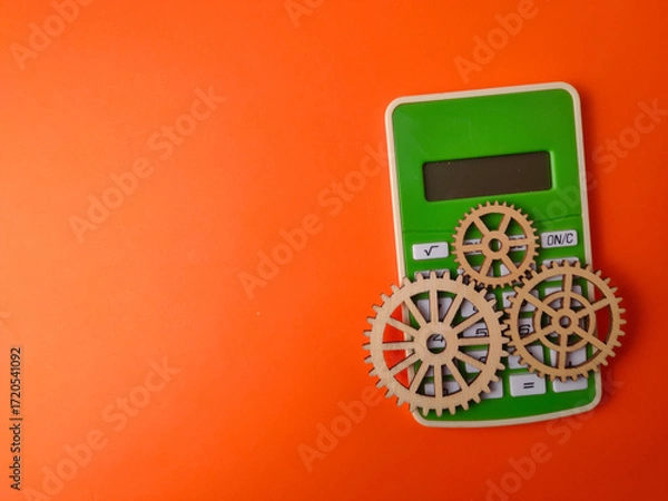 Fototapeta The image displays a green calculator accompanied by three wooden gears on an orange background