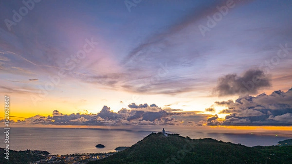 Fototapeta Aerial hyper lapse view of A breathtaking sunset paints the sky in vivid hues of orange and blue over a coastal town, with lush green hills framing the scene. cloud in dramatic sunset over the ocean