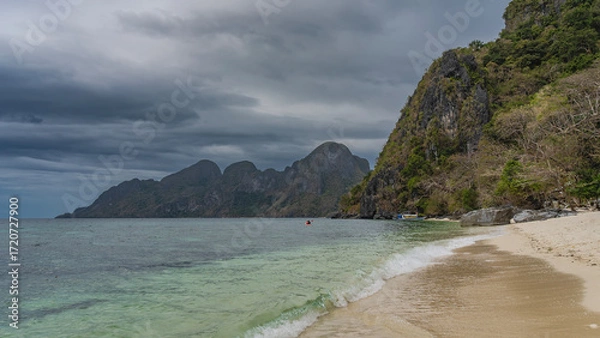 Fototapeta A beautiful tropical beach without people. The waves of the turquoise ocean foam and spread across the sandy beach. Boulders near the water. Bangka boat is moored far away. Mountains, blue sky, clouds