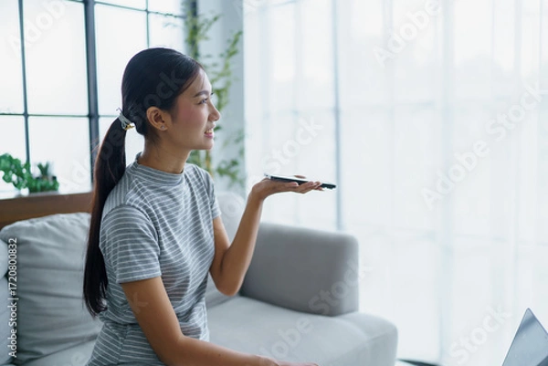 Fototapeta Asian woman sitting on sofa and holding smartphone near mouth talking with AI. using voice assistance at home