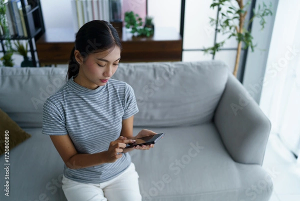 Fototapeta Asian women wear casual cloth texting messaging on smartphone while sitting on sofa at home