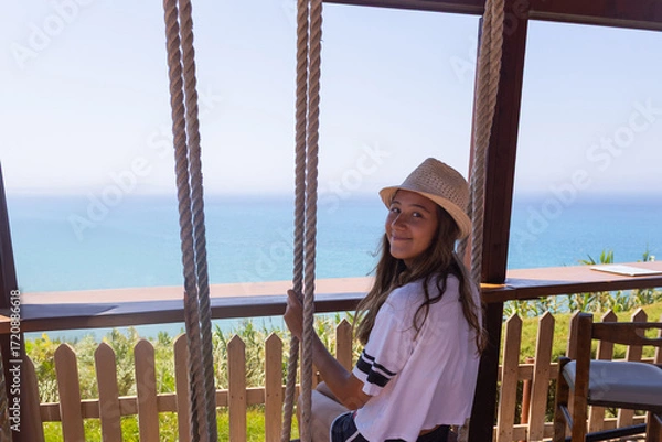 Fototapeta Smiling girl in a sun hat sits on a wooden swing, looking back at the camera with a scenic ocean view behind her