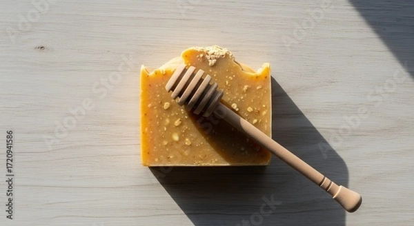 Fototapeta Handcrafted honey soap bar with a wooden honey dipper on a rustic light wood surface, showcasing natural skincare and artisanal products.