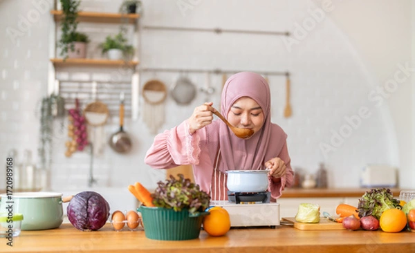 Fototapeta Muslim woman happy cooking healthy meal in kitchen wearing hijab making soup in pot with fresh vegetables food preparation activity nutrition, ramadan healthy recipe preparation, islamic food culture