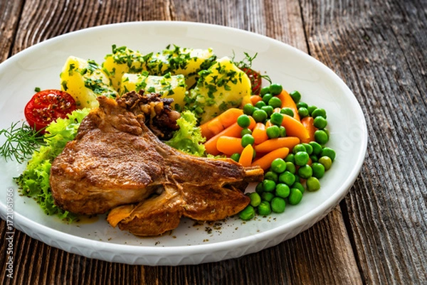 Fototapeta Baked veal cutlet with bone, boiled potatoes, mini carrots and green peas on wooden table