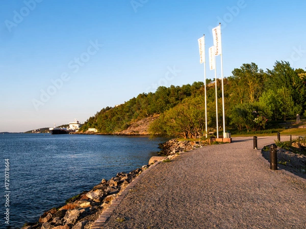 Fototapeta Scenic Waterfront Pathway with Flags and Green Hillside
