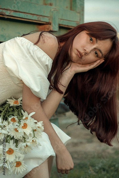 Fototapeta Thoughtful young woman with daisies sitting by farm trailer.Retro grain effect