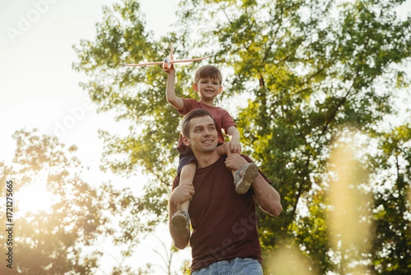 Fototapeta Cheerful father carries his joyful son on his shoulders in park at sunset. Boy is holding a toy airplane and smiling as play with dad outdoors. Child care and happy fatherhood concept. Space for text