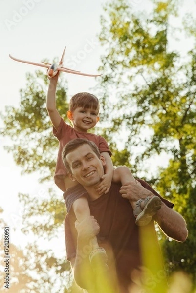 Fototapeta Excited young boy midair, smiling joyfully while being tossed up by a parent on a sunny day with blue sky and trees. POV view, a father high throws and catches laughing son in a park. Vertical video