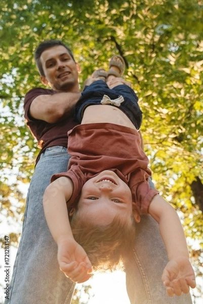 Fototapeta Playful dad holds his son upside down in a sunny park, as the boy laughs joyfully surrounded by green trees and sunlight. Young father having fun while playing with his son outdoors.