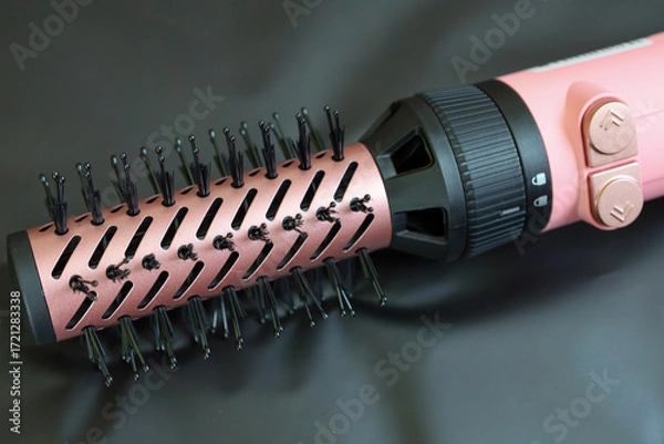Fototapeta ​A macro photograph shows a pink attachment for a hot air brush, with plastic bristles visible on a mesh cylindrical surface. The attachment mechanism is shown in detail, lying on a dark background.