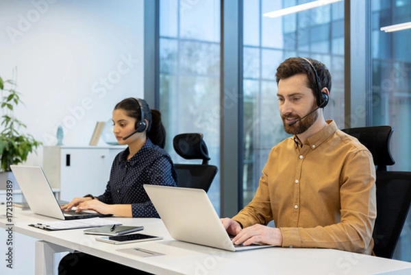 Fototapeta Two diverse call center operators wearing headsets and using laptops while working at desks in a bright, contemporary open-plan office, providing efficient telemarketing and online technical support