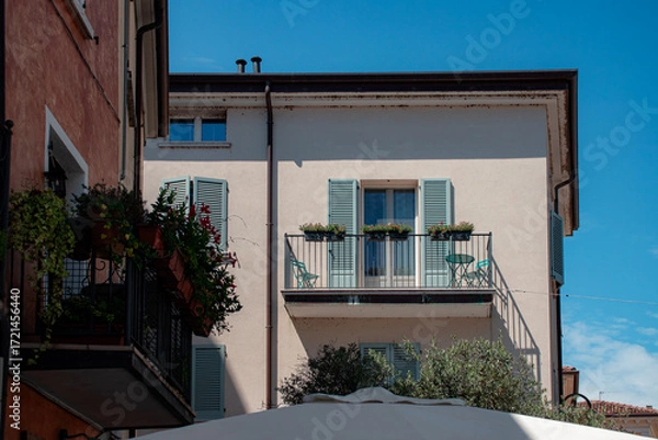 Fototapeta Charming European-style balcony featuring classic wooden window shutters, a black wrought iron railing adorned with potted flowers, and a small cozy bistro seating area
