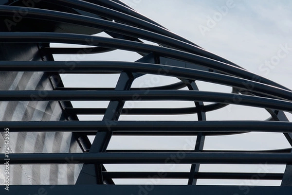 Fototapeta Close-up of a black metal architectural structure. The interlocking curved and straight bars create a dynamic geometric pattern against a pale blue sky.