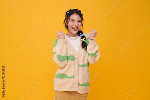 Fototapeta Smiling Asian woman with clenched fists in casual fashion outfit expressing excitement positivity confidence motivation and joyful lifestyle isolated on yellow background