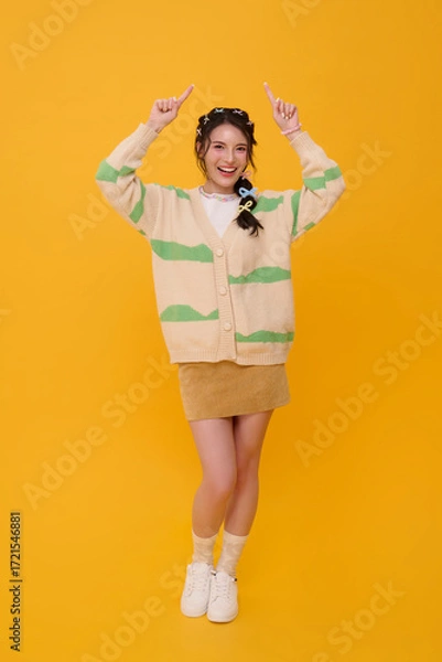 Fototapeta Smiling Asian woman pointing finger upward with both hands in casual fashion outfit expressing positivity confidence joyful lifestyle isolated on yellow copy space background