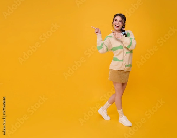 Fototapeta Cheerful Asian woman pointing finger with playful expression in casual fashion outfit standing with fun pose showing positivity confidence and lifestyle isolated on yellow copy space background