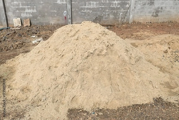 Fototapeta A large, loose pile of light-colored sand sits on the ground in front of a gray cinder block wall at a construction or work site.