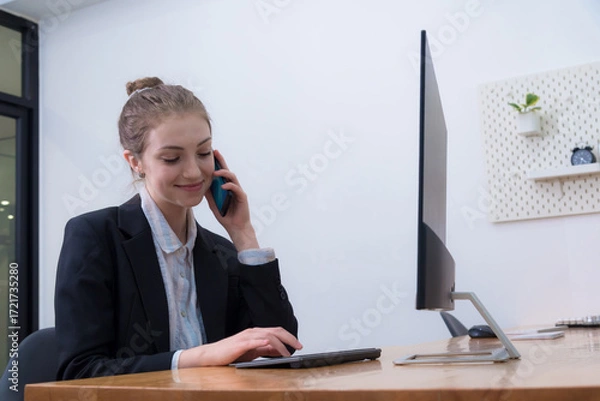Fototapeta Young professional woman is engaged phone conversation while using tablet her desk modern office environment. setting is minimalistic, featuring computer monitor and decorative elements.