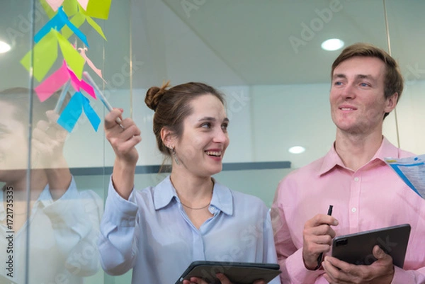 Fototapeta Collaborative team meeting in modern office setting, featuring professionals engaged in brainstorming with colorful sticky notes on glass wall. atmosphere is dynamic and focused.