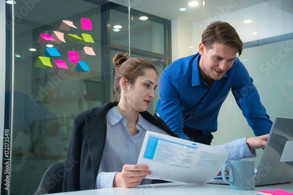 Fototapeta Efficient teamwork in modern office setting showcases woman reviewing documents while man assists her with laptop. Colorful sticky notes in background add creative touch