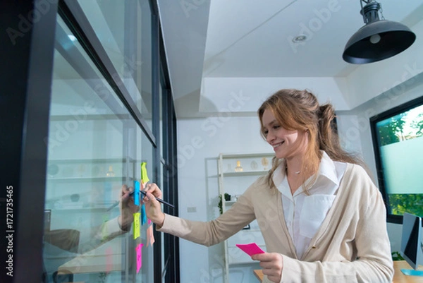 Fototapeta Creative professional engaged in brainstorming session, using colorful sticky notes on glass wall in modern office environment. Focused expression reflects determination and innovation