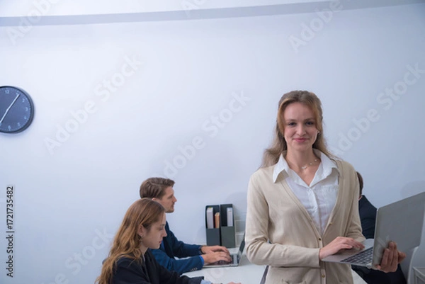 Fototapeta Confident woman stands in modern office, holding laptop, while colleagues work at table in background. atmosphere is professional and collaborative, showcasing teamwork and productivity
