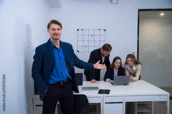 Fototapeta Confident man in blue blazer stands in office, gesturing towards group of colleagues engaged with laptop. atmosphere is collaborative and professional, showcasing teamwork and productivity