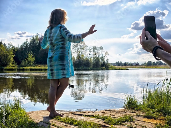Fototapeta Woman enjoying a sunny day by the river while another person captures the moment on a phone