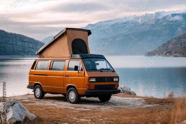 Fototapeta Vintage orange camper van parked by serene lake with mountains in the background, showcasing outdoor adventure and travel lifestyle in a tranquil setting
