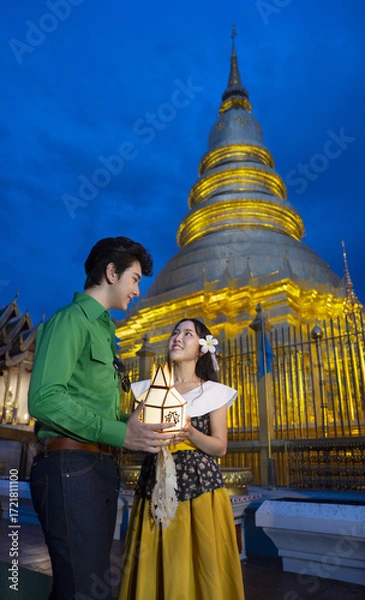 Fototapeta Couple in traditional attire holds lantern in front of illuminated temple at dusk, creating serene and cultural atmosphere