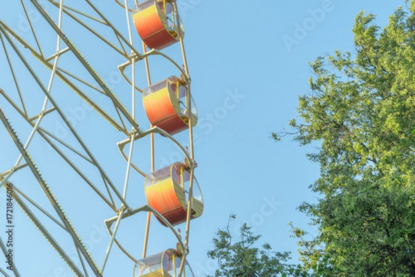 Obraz Ferris Wheel And Green Trees Against Blue Sky. Summer Amusement Park Scene With Ferris Wheel Rising Above Lush Foliage And Sunlight.