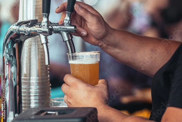 Obraz Close-up of a bartender pouring fresh draft beer into a plastic cup at an outdoor festival. Cold foamy lager being served from a tap, symbolizing refreshment, brewing and celebration