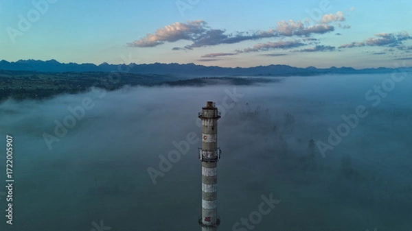 Fototapeta Abandoned industrial chimney tower emerging through morning fog with mountain landscape