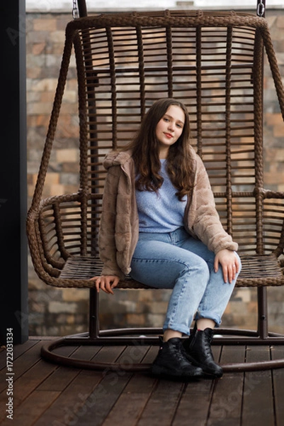 Fototapeta A girl in blue jeans and a warm jacket sits on a wicker bench on the terrace