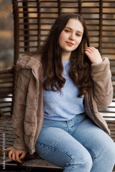 Fototapeta A girl in blue jeans and a warm jacket sits on a wicker bench on the terrace