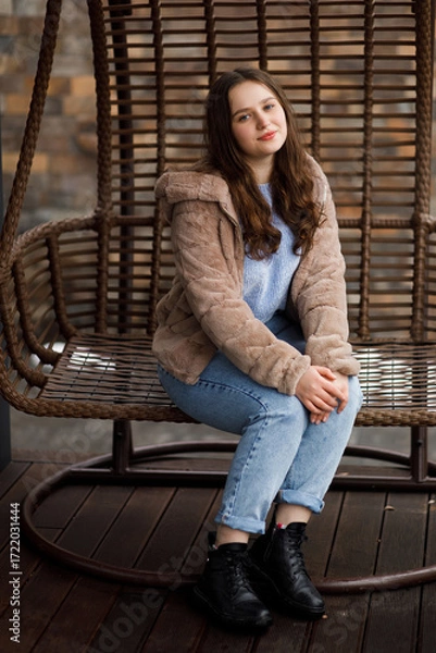 Fototapeta A girl in blue jeans and a warm jacket sits on a wicker bench on the terrace