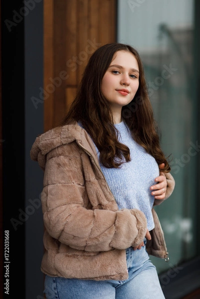 Fototapeta A girl in blue jeans and a warm jacket against the background of a bench.