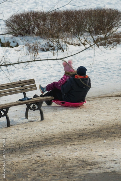 Obraz A man and his daughter enjoy sliding down the snow on a snowboard in a park on a winter afternoon