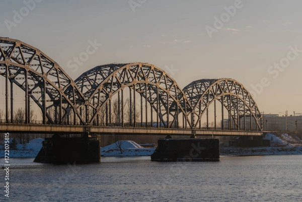 Obraz Sunset landscape of an iron-framed arched bridge crossing a river in a snowy winter