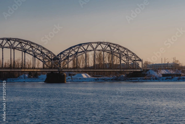 Fototapeta Sunset landscape of an iron-framed arched bridge crossing a river in a snowy winter