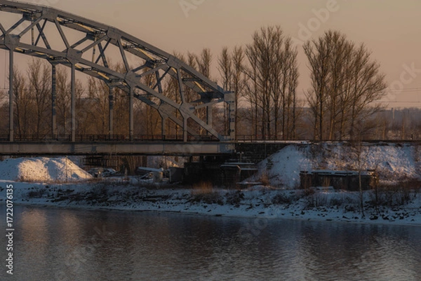 Obraz Sunset landscape of an iron-framed arched bridge crossing a river in a snowy winter